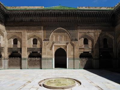 A sunlit interior courtyard of a historic Islamic school featuring intricate stucco carvings, a central horseshoe archway, green-tiled lower walls, and a circular stone basin in the foreground.