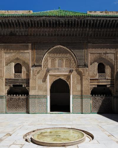 A sunlit interior courtyard of a historic Islamic school featuring intricate stucco carvings, a central horseshoe archway, green-tiled lower walls, and a circular stone basin in the foreground.