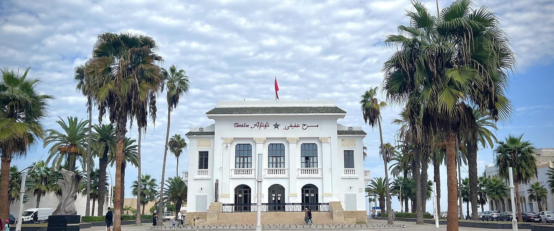 A white neoclassical building with arched windows and balconies, fronted by palm trees and a paved plaza, under a cloudy sky; signage above the entrance reads "Théâtre Afifi" in French and Arabic script, with a red Moroccan flag on the roof.