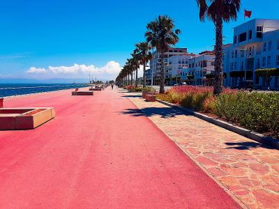 A wide, bright red paved promenade runs along a coastline with palm trees, benches, and multi-story buildings on the right, under a clear blue sky with a Moroccan flag visible on a building.