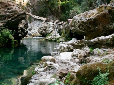 A rustic wooden plank bridge spanning a clear, turquoise river gorge surrounded by rocky cliffs and dense green vegetation.