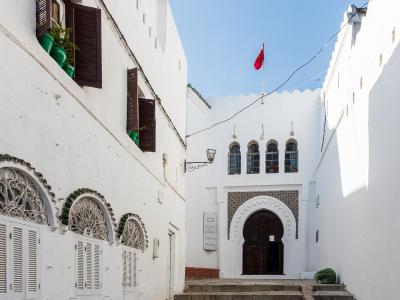 A white-walled courtyard with Moorish-style arched doorways, decorative tilework, and a Moroccan flag flying above the entrance to a building labeled "MUSÉE DE LA KASBAH".