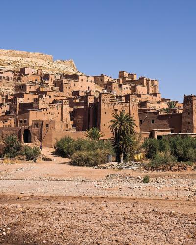A wide-angle photograph of a large, fortified earthen village (ksar) built into a hillside, with palm trees and a dry riverbed in the foreground.