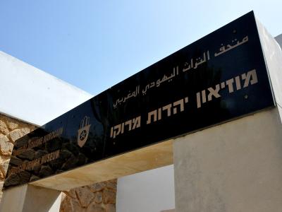A close-up of a black entrance sign for a museum featuring text in Arabic, Hebrew, and French/English, set against a white building with stone cladding.