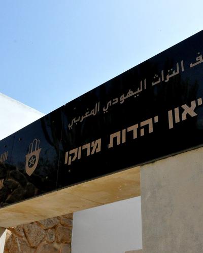 A close-up of a black entrance sign for a museum featuring text in Arabic, Hebrew, and French/English, set against a white building with stone cladding.