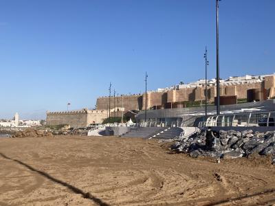 A sandy beach in the foreground with a modern amphitheater-style seating area, tall light poles, and a large historic stone fortress wall (Kasbah of the Udayas) rising above it under a clear blue sky.