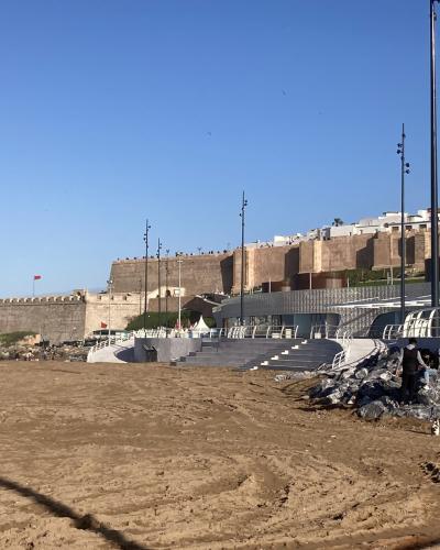 A sandy beach in the foreground with a modern amphitheater-style seating area, tall light poles, and a large historic stone fortress wall (Kasbah of the Udayas) rising above it under a clear blue sky.