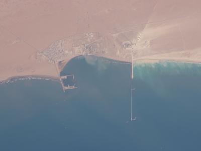 An orbital satellite image showing a coastal settlement with a grid-like street pattern, a small harbor structure, and a long pier extending into the ocean, surrounded by desert terrain.