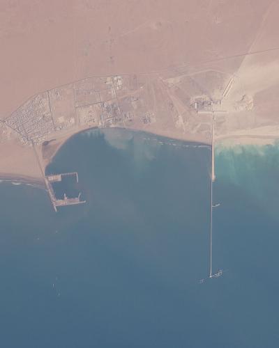 An orbital satellite image showing a coastal settlement with a grid-like street pattern, a small harbor structure, and a long pier extending into the ocean, surrounded by desert terrain.