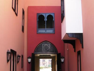 A narrow, covered courtyard or entrance hallway with pink and red walls, arched doorways, and traditional Moroccan-style windows and lanterns.