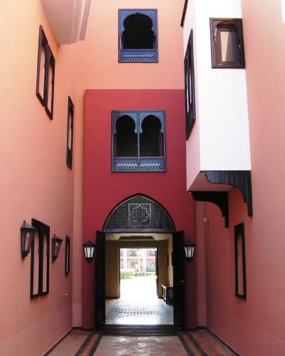 A narrow, covered courtyard or entrance hallway with pink and red walls, arched doorways, and traditional Moroccan-style windows and lanterns.