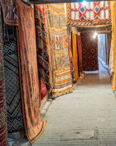 A narrow, brick-paved alleyway within a covered market, lined on both sides with numerous colorful, patterned carpets and rugs hanging from the walls and ceiling.