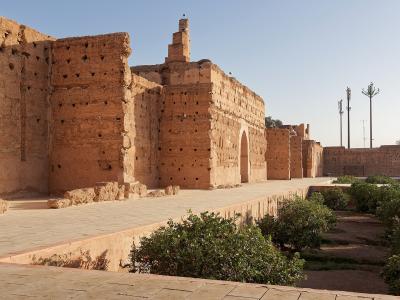 A view of the sunlit, terracotta-colored earthen ruins of a large historical palace complex, featuring high walls with distinctive wooden beam holes, a paved terrace, and a lower garden area with citrus trees.