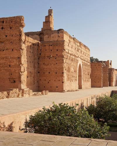 A view of the sunlit, terracotta-colored earthen ruins of a large historical palace complex, featuring high walls with distinctive wooden beam holes, a paved terrace, and a lower garden area with citrus trees.