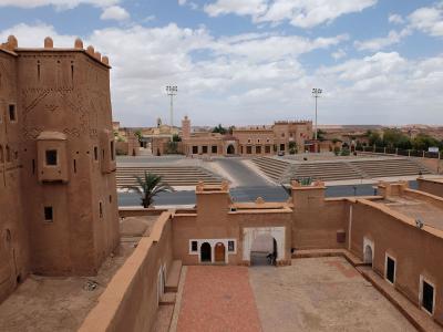 A view from an elevated terrace of a large, traditional mud-brick (pisé) fortified complex with decorative geometric patterns on the walls, overlooking a paved plaza with tiered seating and modern floodlights in the background.