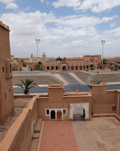 A view from an elevated terrace of a large, traditional mud-brick (pisé) fortified complex with decorative geometric patterns on the walls, overlooking a paved plaza with tiered seating and modern floodlights in the background.