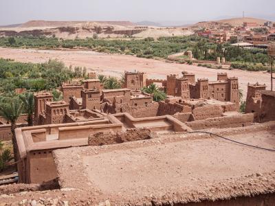 A high-angle view of a traditional rammed-earth fortified village (ksar) with distinctive towers and flat roofs, situated next to a dry riverbed and surrounded by palm groves and arid hills.