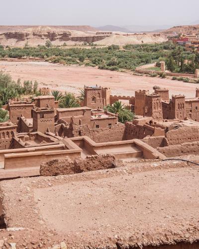 A high-angle view of a traditional rammed-earth fortified village (ksar) with distinctive towers and flat roofs, situated next to a dry riverbed and surrounded by palm groves and arid hills.