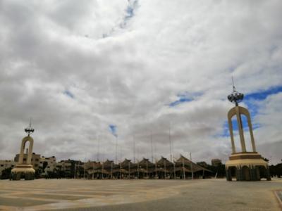 A wide paved public square featuring two tall, identical beige archway monuments topped with decorative metal spheres, with a row of tent-like structures and flagpoles in the background under a cloudy sky.