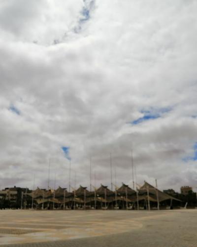 A wide paved public square featuring two tall, identical beige archway monuments topped with decorative metal spheres, with a row of tent-like structures and flagpoles in the background under a cloudy sky.
