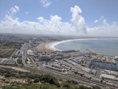 A high-angle panoramic view of a coastal city featuring a long curved beach, a large marina with breakwaters, and a dense urban area extending inland under a partly cloudy sky.
