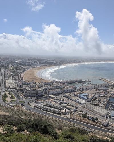 A high-angle panoramic view of a coastal city featuring a long curved beach, a large marina with breakwaters, and a dense urban area extending inland under a partly cloudy sky.
