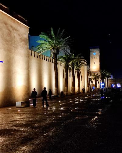 A night scene showing a long, illuminated stone rampart wall with crenellations, a row of palm trees, and a clock tower in the background, with people walking on a wet paved area.