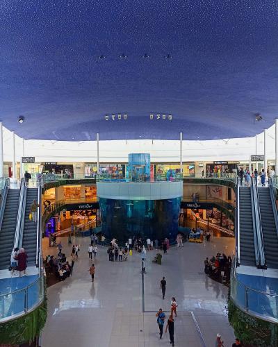 The image shows the interior atrium of a large shopping mall featuring a massive cylindrical aquarium in the center, surrounded by multiple levels of walkways and escalators, with a dark blue ceiling dotted with fiber optic lights resembling a starry