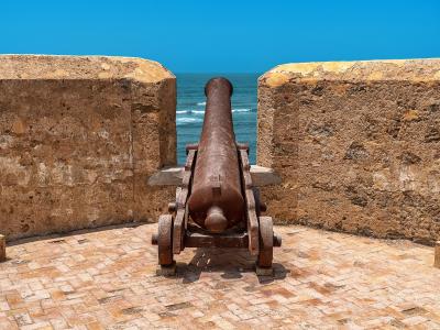 An old, rusty cannon mounted on a wooden carriage sits on a brick-paved terrace between two weathered stone walls, overlooking the blue ocean.