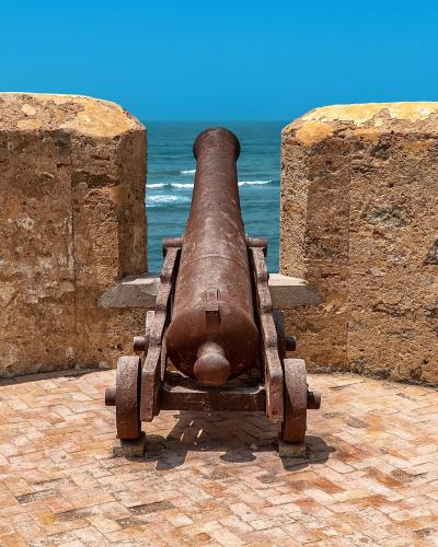 An old, rusty cannon mounted on a wooden carriage sits on a brick-paved terrace between two weathered stone walls, overlooking the blue ocean.