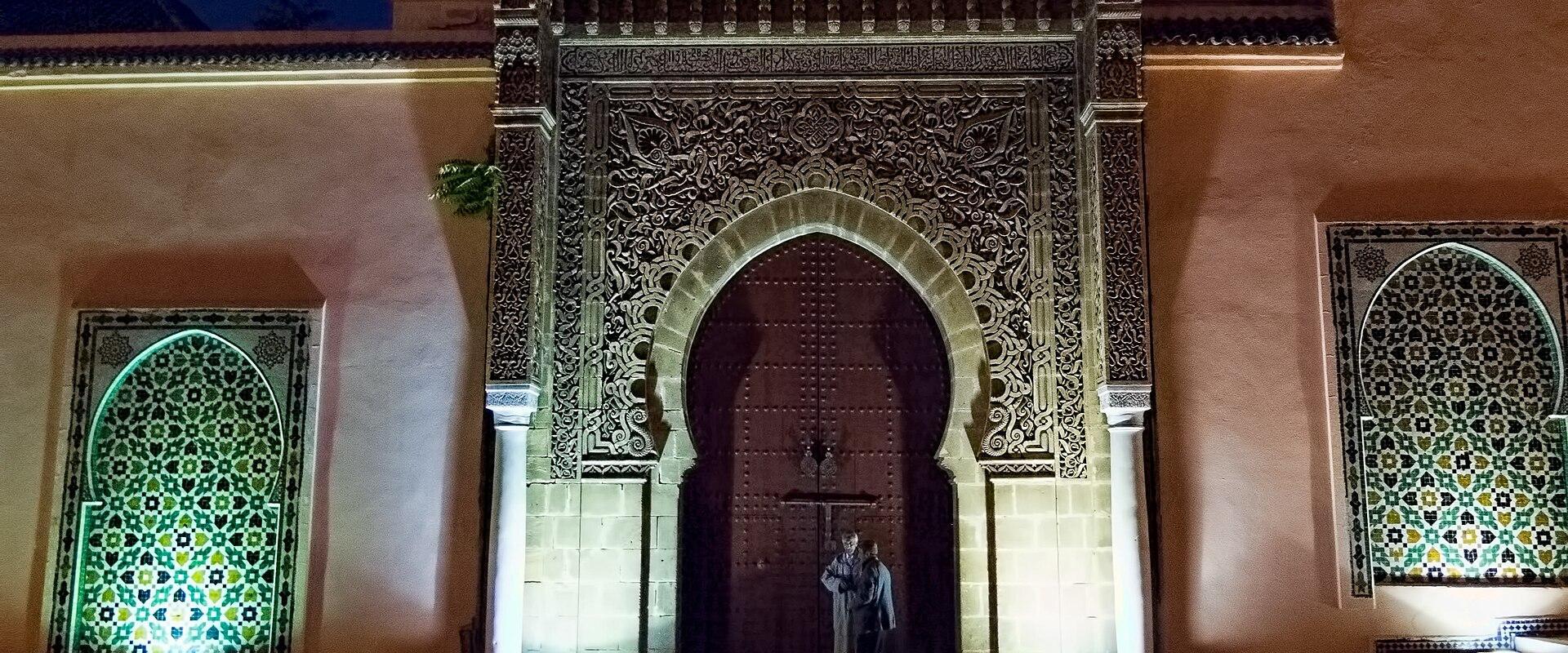 A large, ornately carved wooden door set within a horseshoe archway, flanked by two arched niches with green and yellow zellige tilework, illuminated at night with two people standing in front.